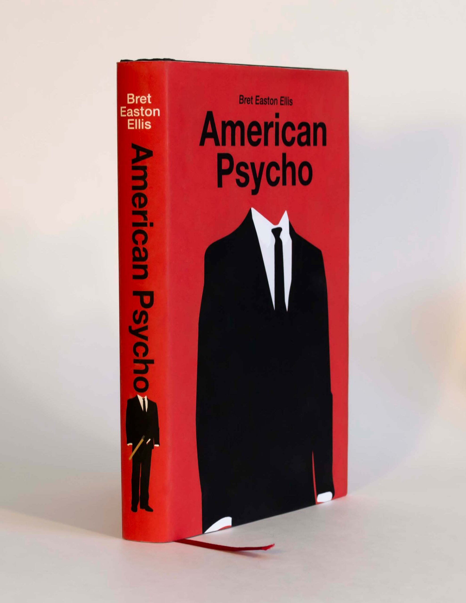 Picture of the book American Psycho standing up with red cover. On the cover there is a minimalist illustration of a suit. The spine of the book has the title and the illustration of the suit holding an axe.